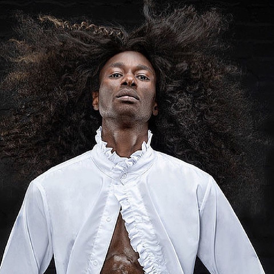 A male model with very long hair being blown and wearing a white ruffled shirt
